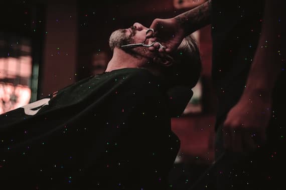 Barber shaving client's beard with razor in dark salon